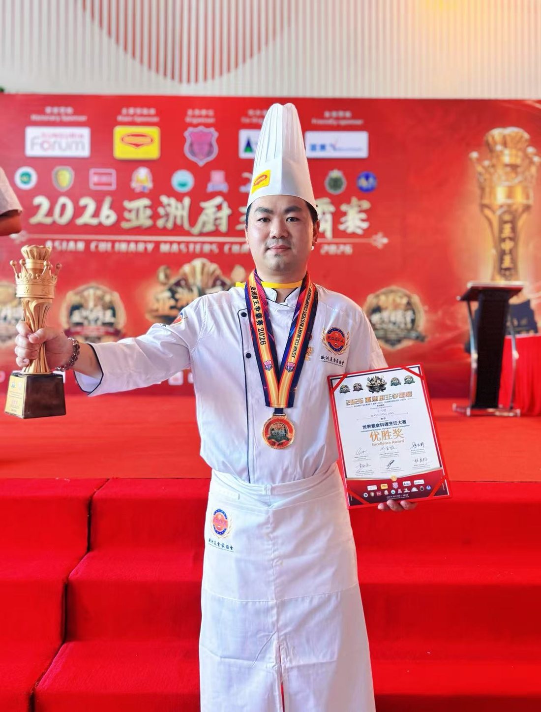 Chef Wang Xing Jian met zijn gouden beker en oorkonde voor de backdrop van de Asian Culinary Masters Championship 2026 in Maleisië